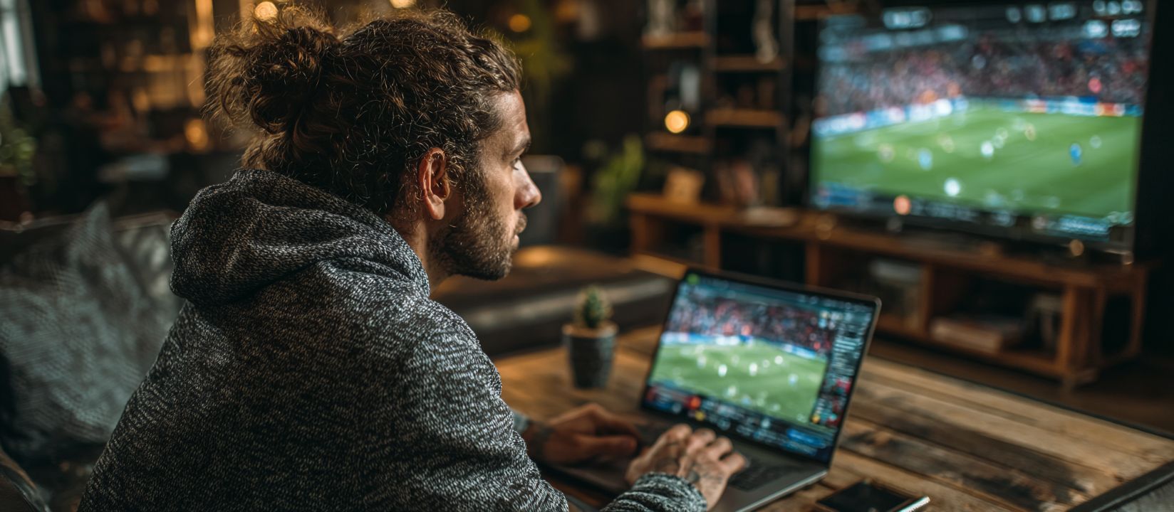 Man betting on live football using laptop while watching match on TV at home - AH444